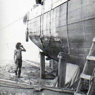 A figure standing beside the wooden hull of a boat supported on the sand.