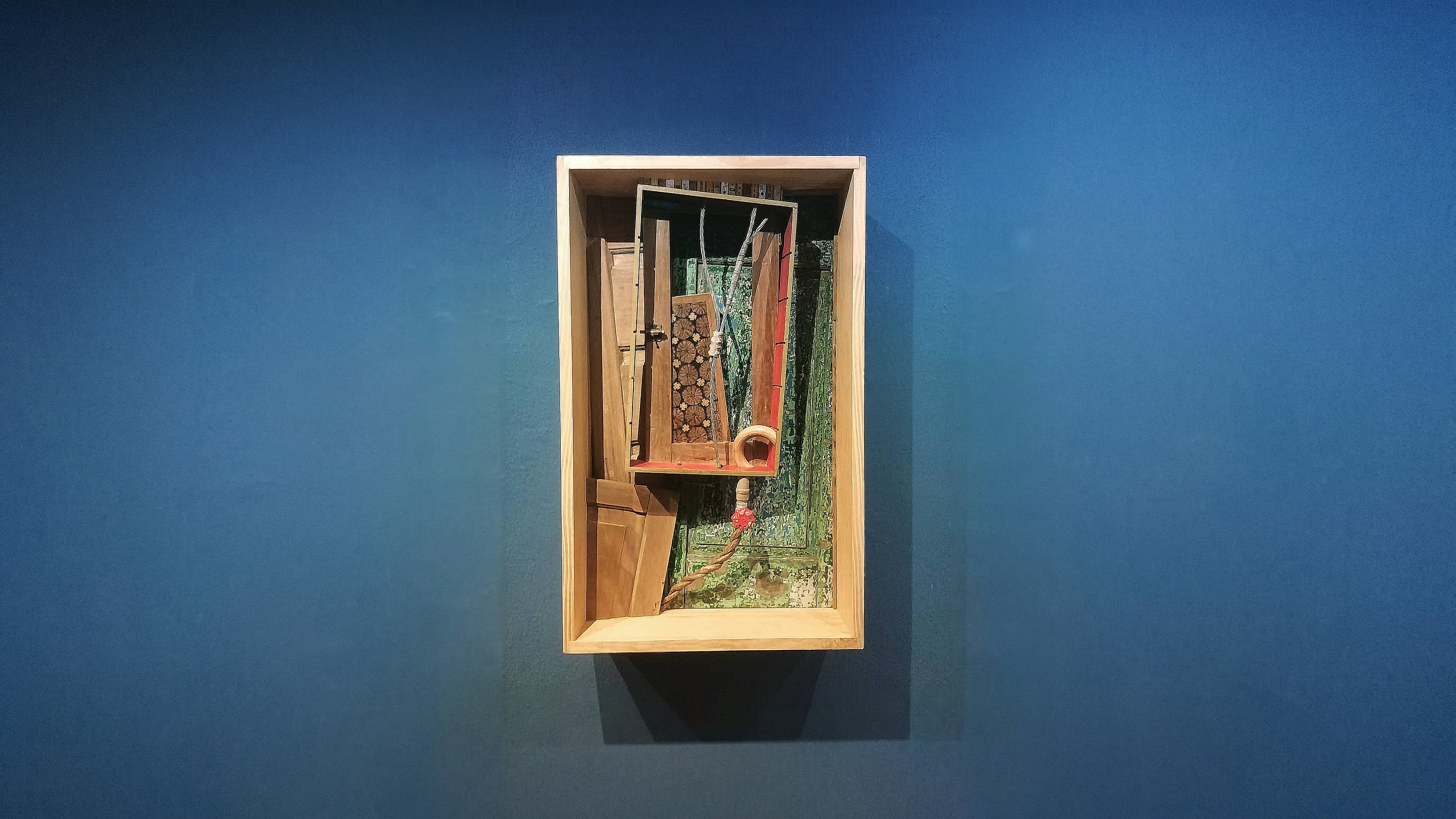 The work Caixa 10 brings together cut wood pieces, painted elements and metal parts against a blue wall.