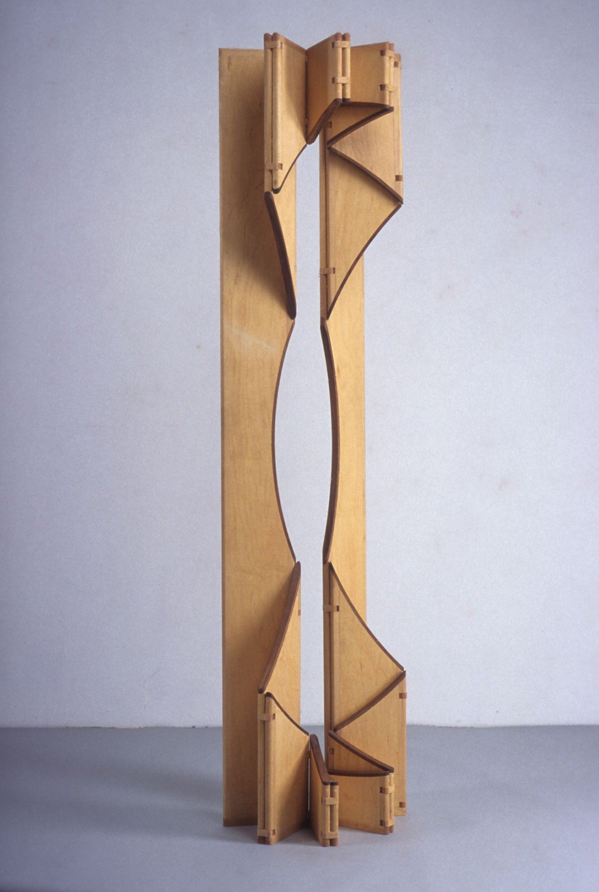 Light wood mobile sculpture with vertical articulated parts and a central opening on a grey floor.