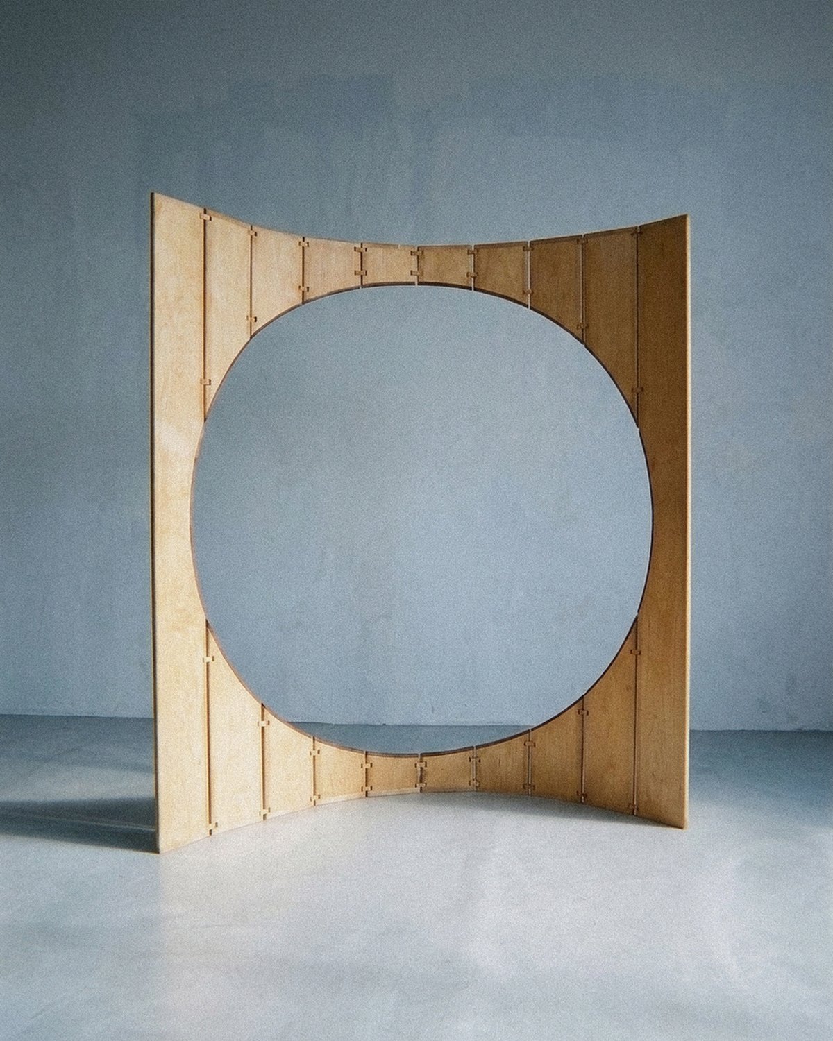 A escultura móvel Ó em madeira e latão forma um arco aberto com grande vão circular sobre piso cinza.