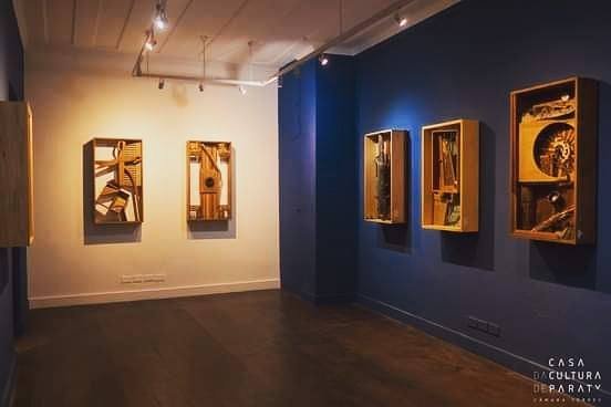 Sala de galeria com caixas de madeira montadas sobre uma parede branca ao fundo e uma parede azul lateral.