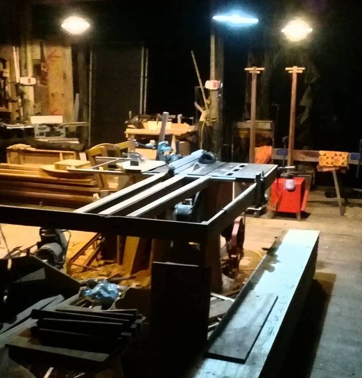 Woodworking workshop at night showing machinery, table saw and timber under overhead lamps.