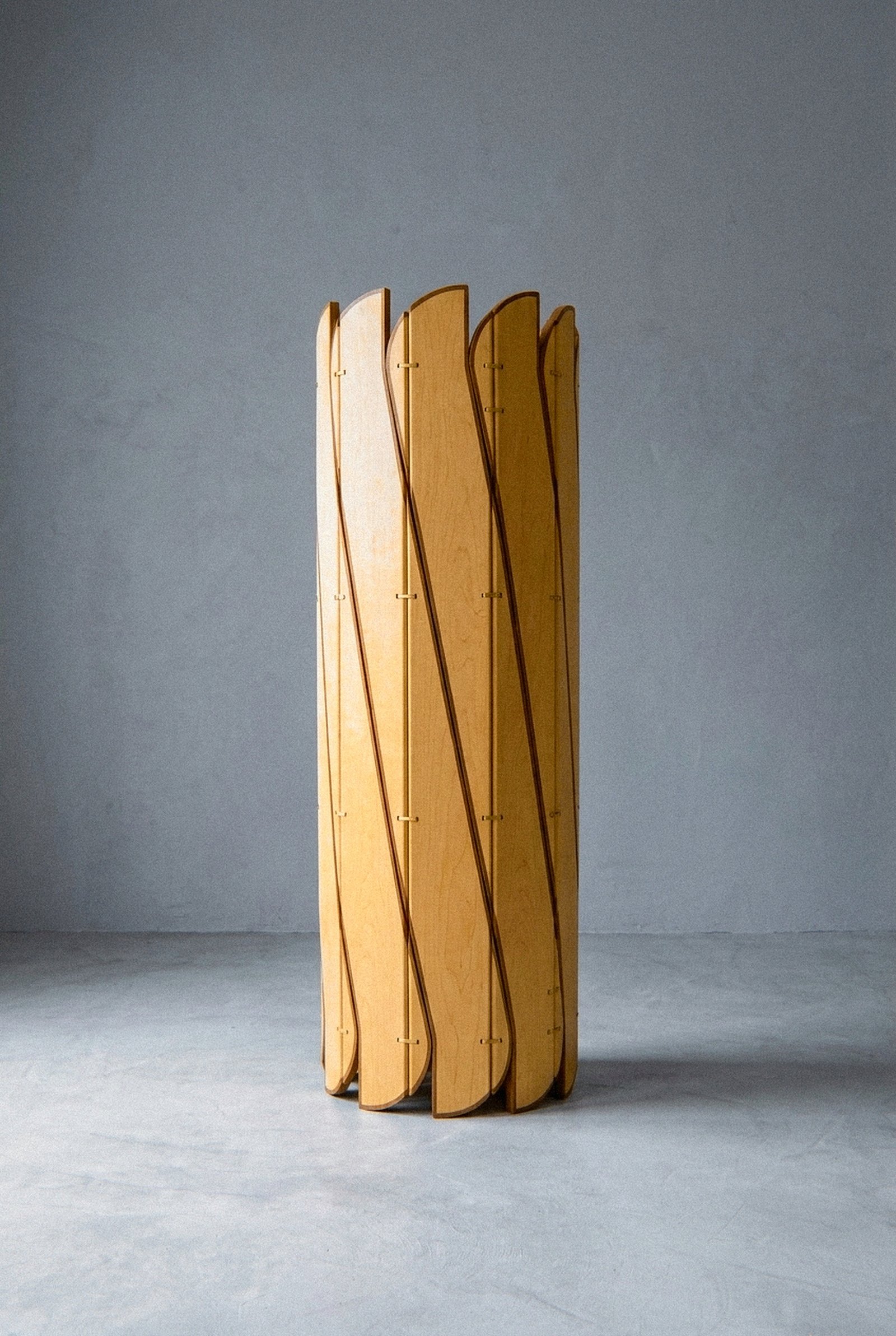 The mobile sculpture Onda in maple plywood, solid ash, and brass stands closed in a cylinder shape on gray flooring.